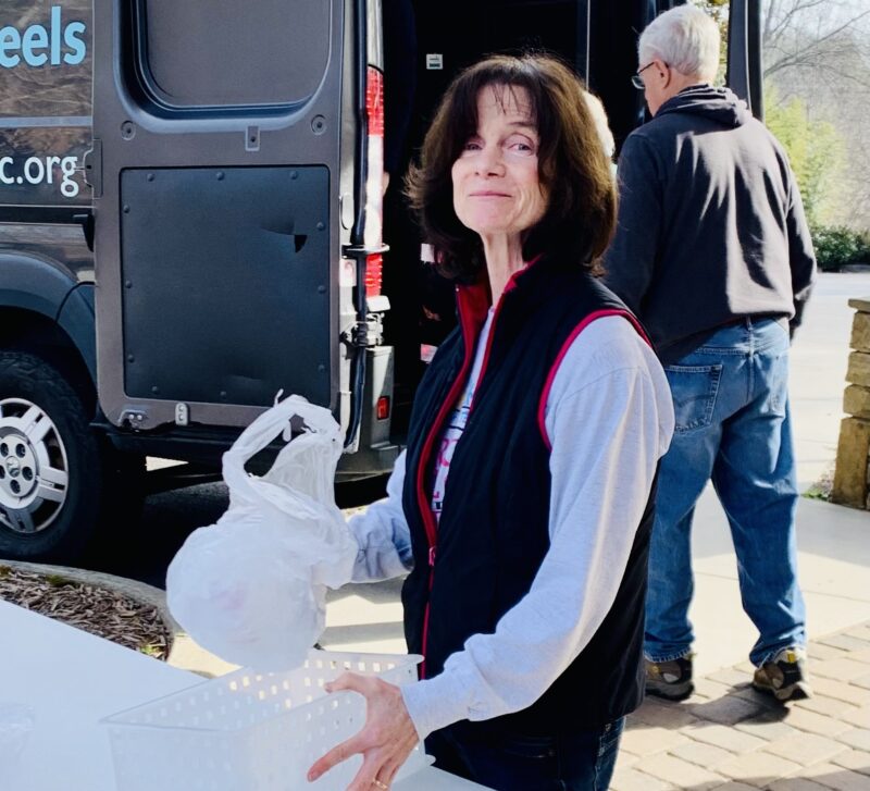 Woman with dark hair wearing a black vest with a long-sleeved white top underneath. She is holding a bag. A man is unloading a van in the background.